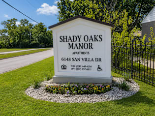 A sign for Shady Oaks Manor Apartments is displayed in front of a fence.