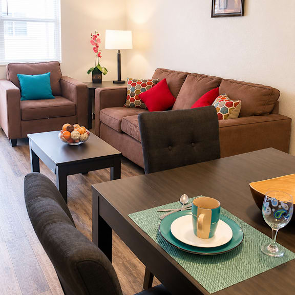 A living room with a brown couch, a brown chair, a brown armchair, a coffee table, a lamp, a vase with flowers, a bowl of fruit, a plate with a cup, and a picture on the wall