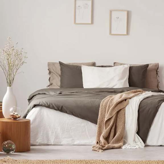 A bed with a grey comforter and pillows is in the center of a room with a white wall and a small table with a vase on it.
