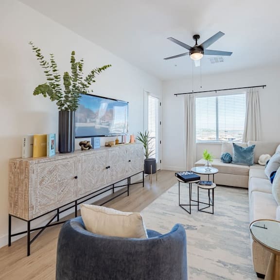 A living room with a grey armchair, a white sofa, a wooden cabinet, a glass table, a TV, a plant, and a ceiling fan.