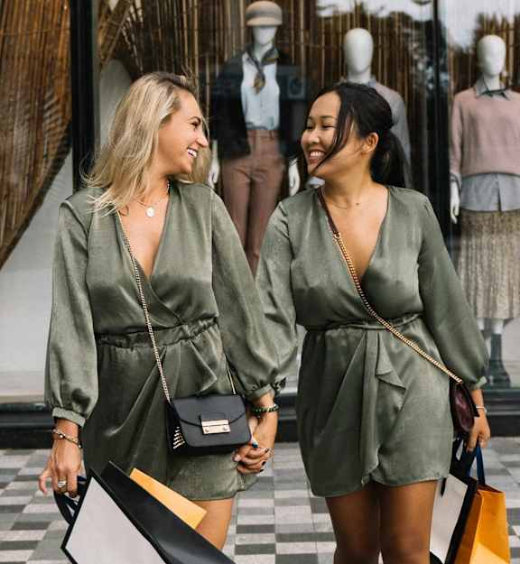 Two women in matching green dresses are walking and talking while holding shopping bags.