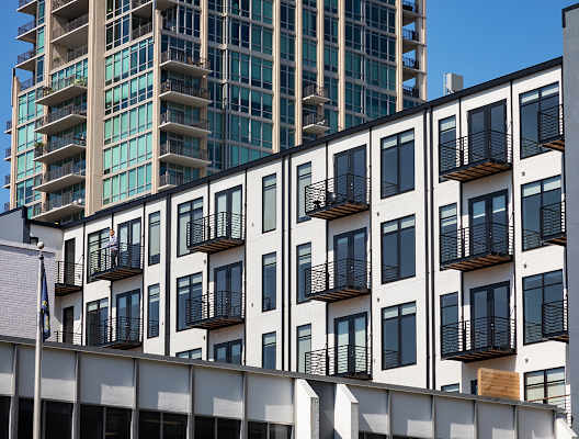 A white building with black balconies is in front of a blue building. at The Elle on Lockwood Apartments, Webster Groves, MO, 63119