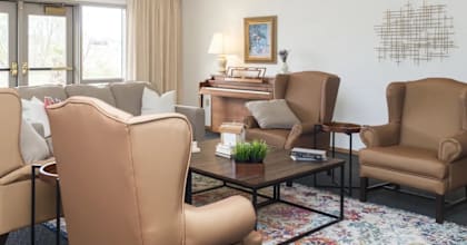 A living room with a brown chair in the foreground and a white wall with a picture frame and a fire alarm.
