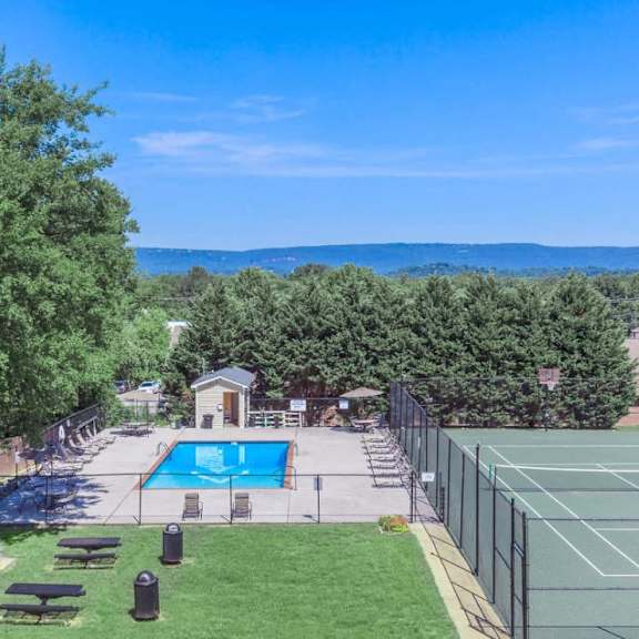 A tennis court is surrounded by a fence and a pool is in the background.