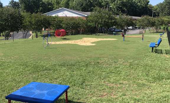 A blue picnic table is in the foreground of a grassy park with a playground and a building in the background.