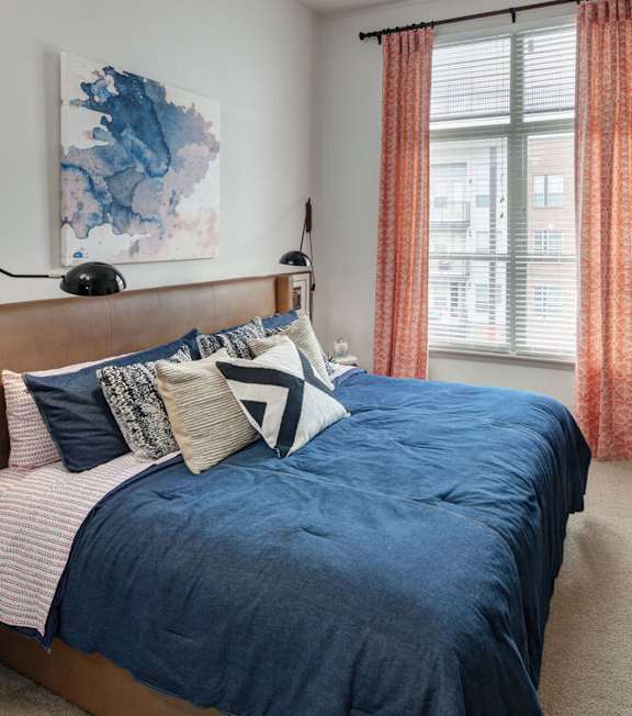A bedroom with a blue bedspread and pillows, a wooden headboard, and a window with orange curtains at Regatta Sloans Lake Apartments, Colorado, 80204