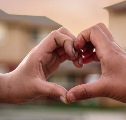 Two hands forming a heart shape against a blurred residential background.