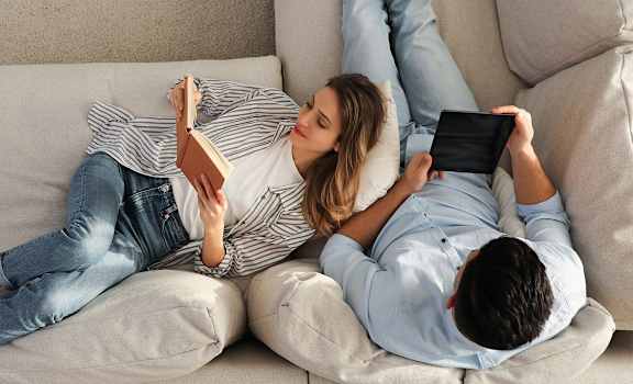 A man and a woman are lying on a couch, the woman is reading a book and the man is using a tablet.