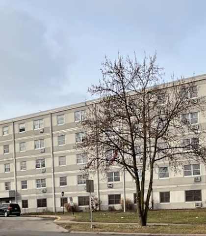 A large, multi-story building with a parking lot in front and trees on the side.