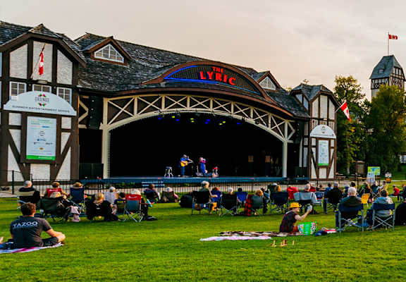 A large crowd of people are sitting on the grass in front of a stage with a sign that says Lyric.