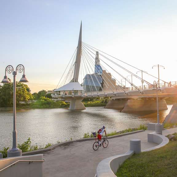 A cyclist rides along a path next to a river with a modern bridge in the background.