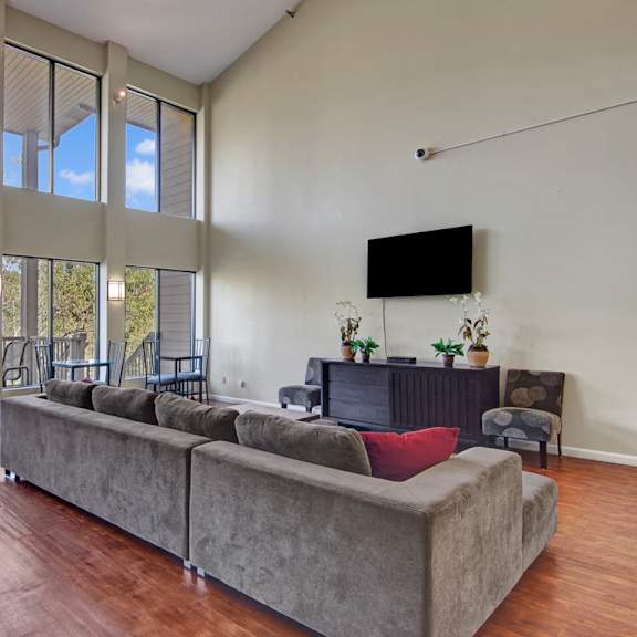 The spacious, modern living room with a couch, throw pillows, large windows, a ceiling fan, wall art, plush chairs, coffee table, decorations, large windows, a TV, and glass doors to the balcony at Town Centre Apartment Homes. 