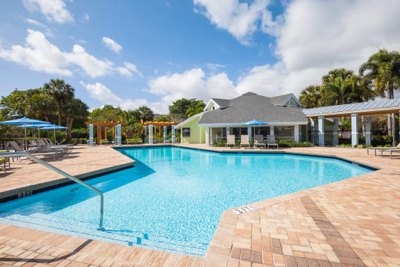 A large swimming pool with a blue tiled edge and a brick patio at The Villages Apartment of Bayan Grove in Boynton Beach, FL