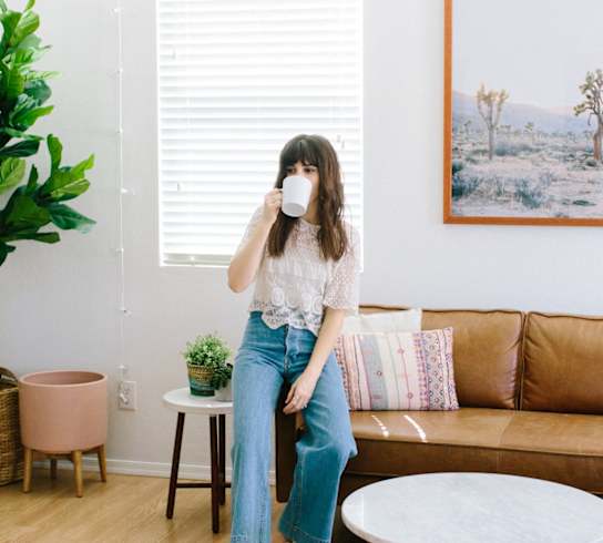 A woman is sitting on a brown couch with a white wall and a painting behind her.