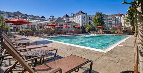 A pool area with sun loungers and umbrellas in front of apartment buildings. at Vermella Lyndhurst apartments, Lyndhurst, NJ, 07071