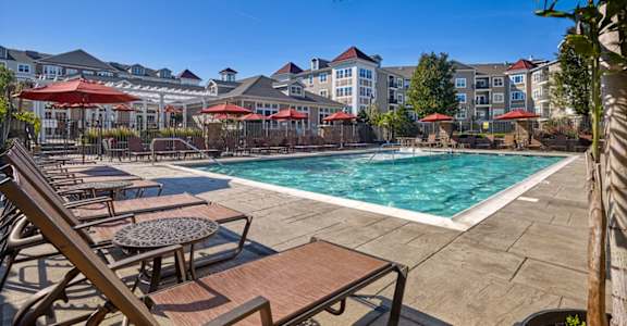 A pool area with sun loungers and umbrellas in front of apartment buildings. at Vermella Lyndhurst apartments, Lyndhurst, NJ, 07071