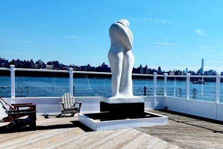 A sculpture of a person holding a ball is on a wooden deck with chairs and a view of the water and city in the background.