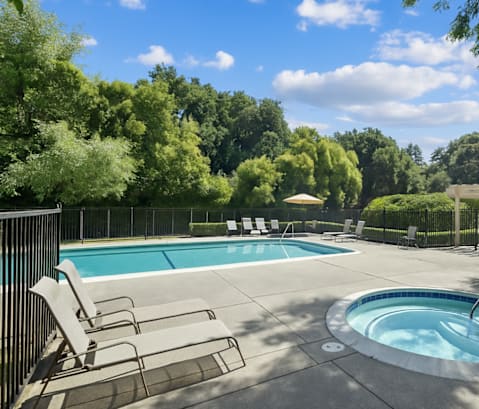 A pool with a hot tub and chairs in the foreground with trees in the background.