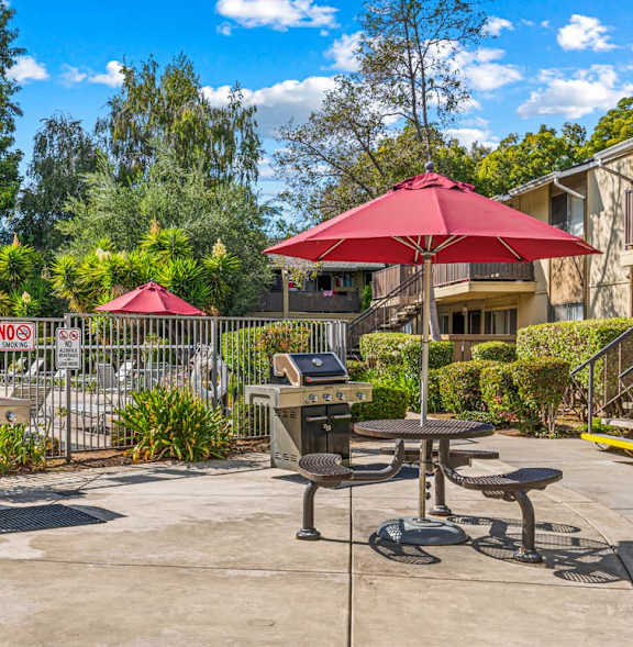 A red umbrella is in the middle of a patio with a picnic table and a grill.