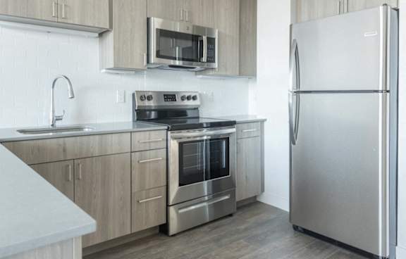 A modern kitchen features light wood cabinets, stainless steel appliances, a gray countertop, and a white subway tile backsplash.