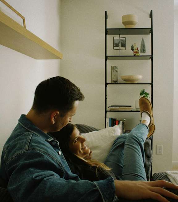 A man and woman are sitting on a couch, with the man wearing a denim jacket and the woman wearing a striped shirt and brown boots.