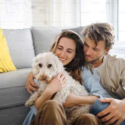 A couple is sitting on a grey couch with a dog between them.