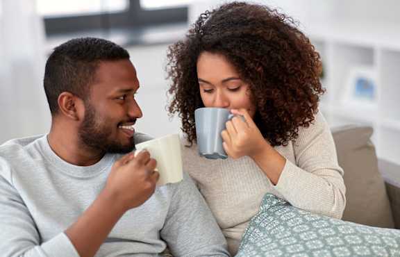 A man and a woman are sitting on a couch, each holding a mug and enjoying a moment together.