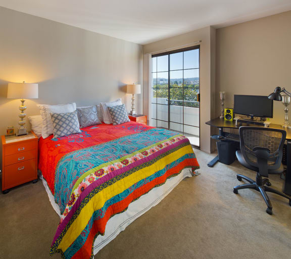 A bedroom with a bed, computer desk, and a view of the balcony.