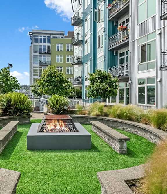 A modern fire pit in the middle of a grassy area surrounded by concrete benches.