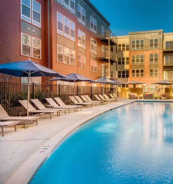A large swimming pool surrounded by lounge chairs and umbrellas in front of a multi-story apartment building.