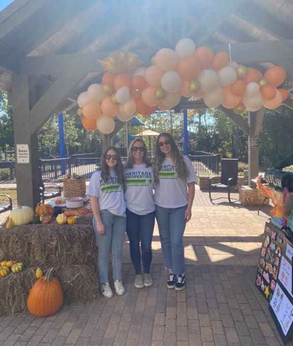 Three women wearing matching t-shirts stand under a balloon arch.