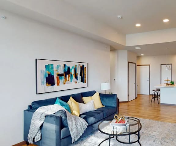 The spacious living room with a blue couch, coffee table, glass coffee table, decorations, a TV, artwork, and view of the dining room with a table, chairs, and place settings for four, and the kitchen at Van Buren West Apartment Homes.