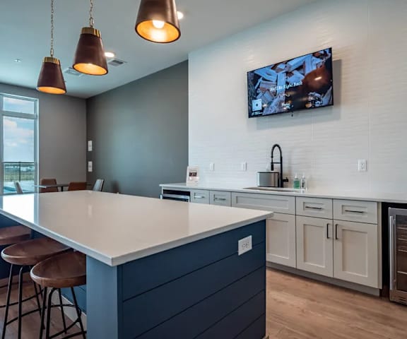 The community kitchen with a fridge, marble counters, overhead lighting, TV, and barstool chairs at Van Buren West Apartment Homes.