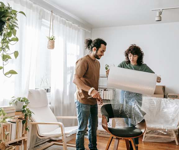 Two people are unrolling a large piece of white material in a room with a white chair and a plant.