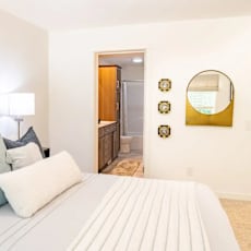 A bed with white and blue pillows is in the foreground of a room with a mirror and a lamp.