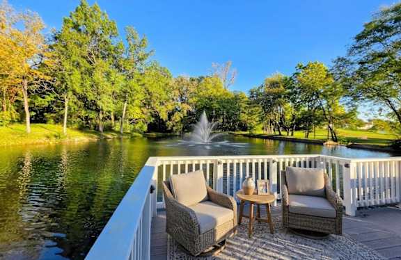 Two chairs and a table are on a deck overlooking a pond.