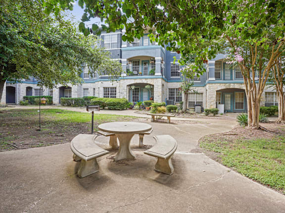 A concrete table and chairs are in the foreground of a courtyard with a building in the background.