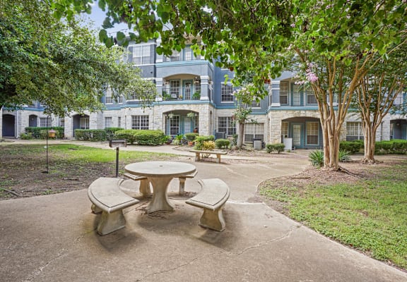 A concrete table and chairs are in the foreground of a courtyard with a building in the background.