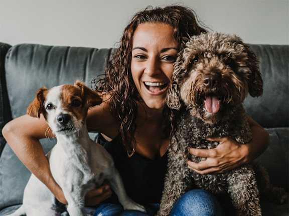 A woman is sitting on a grey couch with two dogs, one white and one brown.
