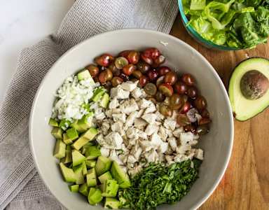 A bowl of food with chicken, avocado, grapes, and greens.