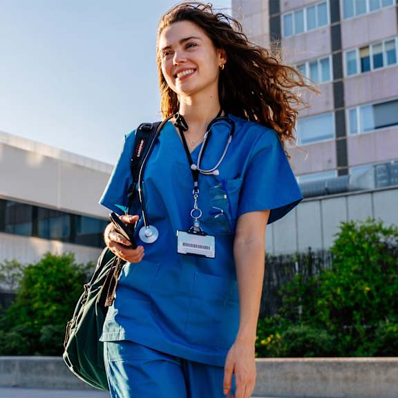 A woman in blue medical scrubs and a stethoscope is walking outside a building.