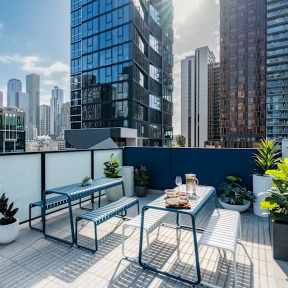 A blue metal table with a white bench on a rooftop terrace with a city skyline in the background.