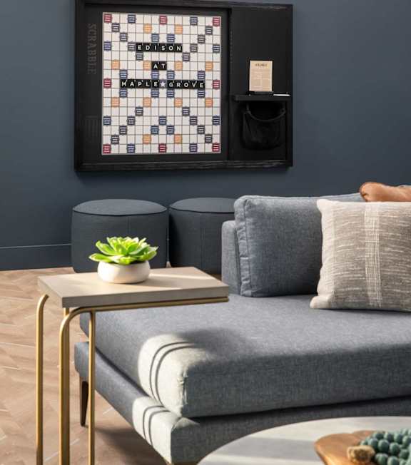 A living room with a grey couch, a small table with a plant and a book, and a framed board game on the wall.at The Edison at Maple Grove, Maple Grove, MN
