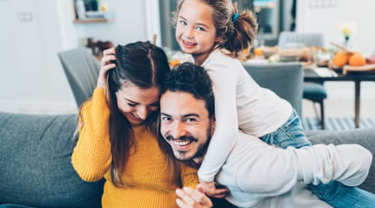 A family of three is sitting on a couch, with the child on the man's shoulders.