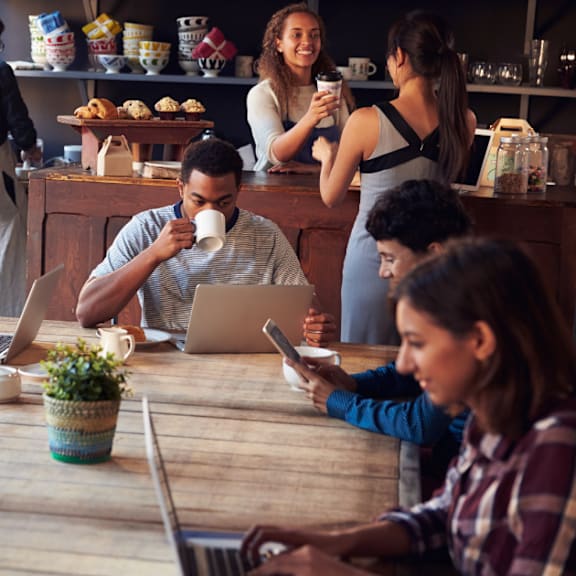 A group of people are sitting around a wooden table in a cafe, using laptops and drinking coffee.