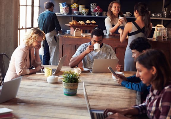 A group of people are sitting around a wooden table in a cafe, using laptops and drinking coffee.
