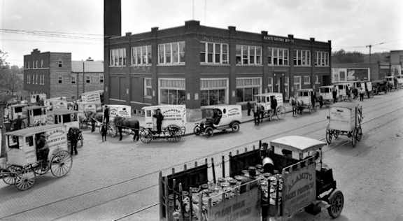 A black and white photo of a street with horse-drawn carriages and a building in the background.