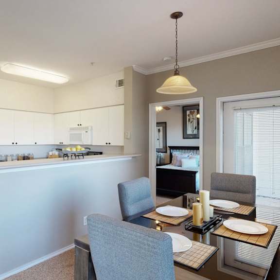 A dining room with a table and gray chairs with the places set with the white kitchen, bathroom, and bedroom in the background at Stoneleigh on May Apartment Homes. 