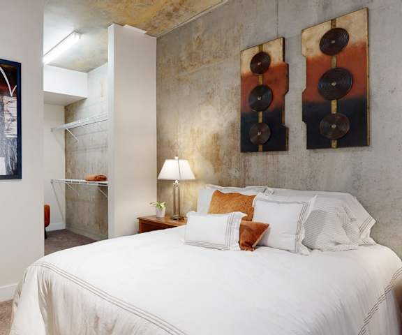 A spacious bedroom with a queen-size bed with a white bedspread, a concrete wall, a dresser, wall art, decorations, closet with shelves, and nightstands at Skyline Lofts Apartment Homes. 