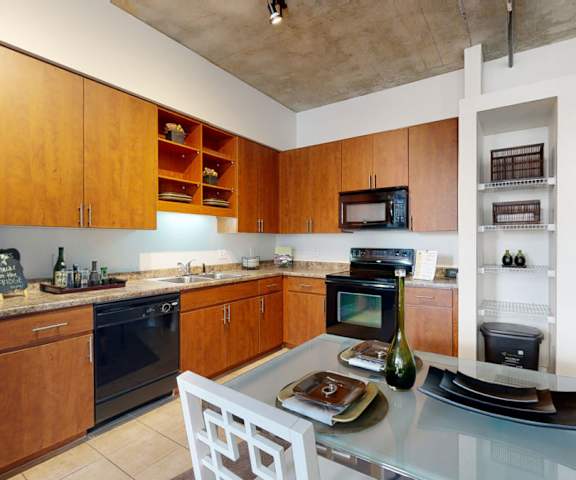 A spacious kitchen with black appliances, a dining table with four places set, marble countertops with lots of room, decorations, wall art, and light wooden cabinets at Skyline Lofts Apartment Homes. 
